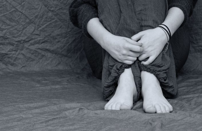 Person som sitter hopkurad med knäna uppdragna mot bröstet och armarna runt benen, barfota på ett grått tygstycke.