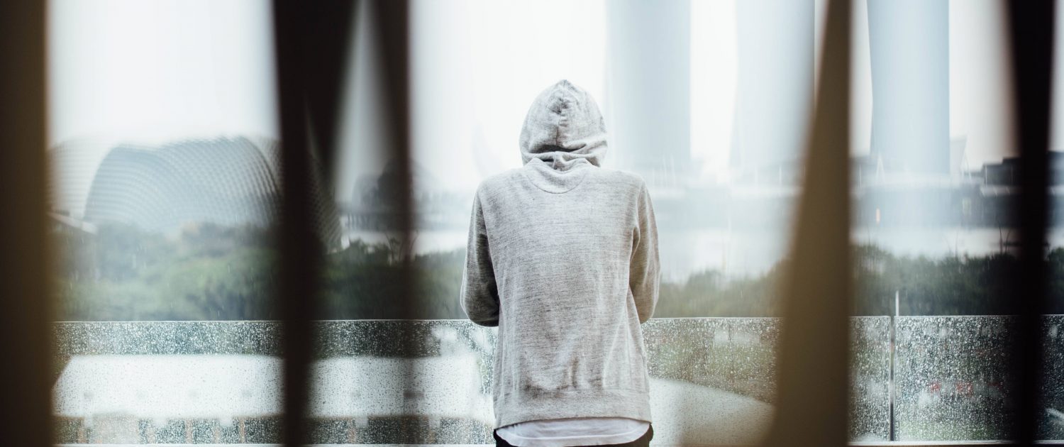Person i grå luvtröja med huvan uppe, sedd bakifrån, som tittar ut över en regnig stad genom ett glasräcke.